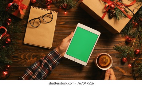 Chrismas flat lay with garlands, gift box, notebook and white tablet computer with green screen. Man using phone and drinking tea. Chroma key. Top view