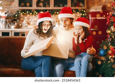 Christams Eve Miracle. Emotional young family opening big shining gift box next to Christmas tree on xmas eve, free space - Powered by Shutterstock