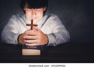 Christian Asian man praying and holding cross on bible. Catholic male praying and worship over bible with beautiful light in dark room.