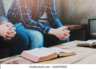 Christian Friends Sitting On A Wooden Chair While Praying Together  With The Holy Bible On Wood Table