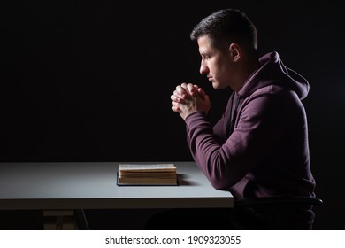 Christian prayer. Man praying before bible. Guy crossed his palms for prayer. He holds his hands over Bible. Young guy praying over black background. He reads Christian Bible. Religious rites