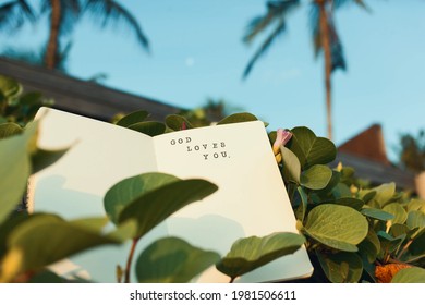 Christian saying 'God loves you' printed with stamps and ink in a diary. Motivational supportive text for religious people in a notebook surrounded by tropical nature, palm trees and greenery