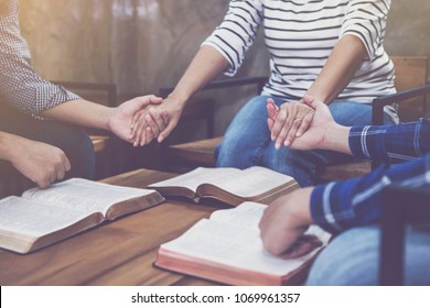Christian small group holding hands and praying together around a wooden table with blurred open bible page in homeroom, devotional or prayer meeting concept