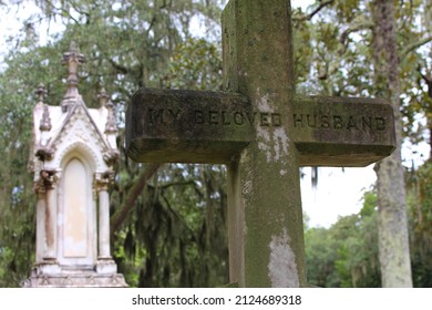 Christian stone cross religious monument to husband and stone structure amidst landscape of Spanish moss trees in outdoor cemetery graveyard.