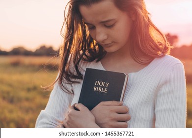 Christian teenage girl holds bible in her hands. Reading the Holy Bible in a field during a beautiful sunset. Concept for faith, spirituality and religion. Peace, hope.