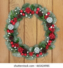 Christmas And Advent Wreath Decoration With Red And Silver Bauble And Gift Box Decorations, Holly, Mistletoe, Pine Cones And Snow Covered Evergreen Blue Spruce Fir On Oak Wood Front Door Background.