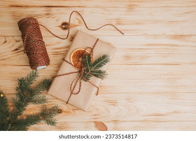 Christmas background. Gift wrapping in brown paper with dry orange and branch of coniferous tree on a light wooden background. The process of packing a Christmas gift. Top view - Powered by Shutterstock