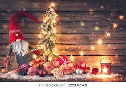 Christmas background. Santa Claus or dwarf holds a fir tree with Christmas lights surrounded by gift boxes and glowing lantern on rustic wooden - Shutterstock ID 1220816908