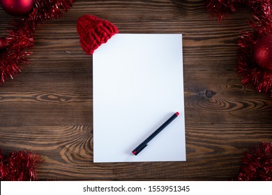 Christmas background. A white sheet of paper with a knitted red hat put on a corner and a pen surrounded by  tinsel, toys and decorations on a brown wooden background. Top view