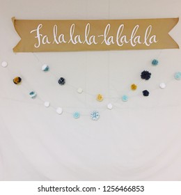 Christmas Banner With Holiday Phrase Written In Script Against A Beige Background. White, Blue, And Gold Pom Pom Garland Strung Underneath The Banner.