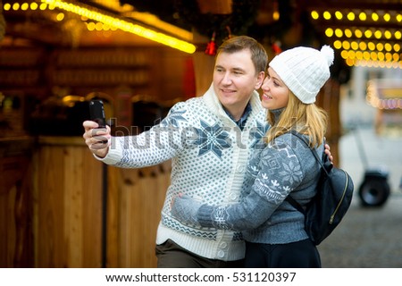 Christmas bazaar. The young couple is photographed near the wooden festively decorated pavilion. A man holds a smart phone in your hand and makes a selfie. Both with pleasure pose. Merry Christmas.