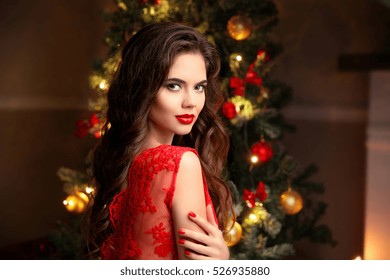 Christmas. Beautiful Smiling Woman. Manicure Nails. Makeup. Healthy Long Hair Style. Elegant Lady In Red Dress Over Christmas Tree Lights Background. Happy New Year.