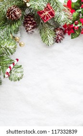 A Christmas Border To Left And Top Of Frame Consisting Of Artificial Foliage, Real Pine Cones And Decorative Ornaments, Sprinkled With Snow On A Fake Snow Background.   Snow Provides Copy Space.