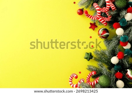 Christmas border with pine branches, candy canes, red and green baubles, stars and pom poms on yellow background. Cheerful festive layout with copy space.