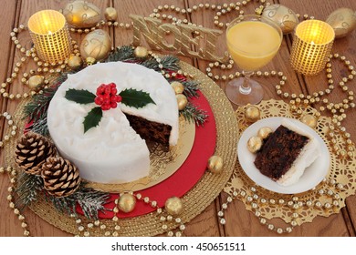 Christmas Cake And Slice, With Egg Nog, Holly Berries, Candles, Noel Glitter Sign, Gold Bauble Decorations And Foil Wrapped Chocolates With Bead Strands Over Oak Table Background.