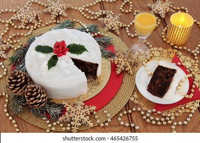 Christmas Cake And Slice, With Egg Nog, Holly Berries, Candles, Gold Bauble Snowflake Decorations With Bead Strands Over Oak Table Background.