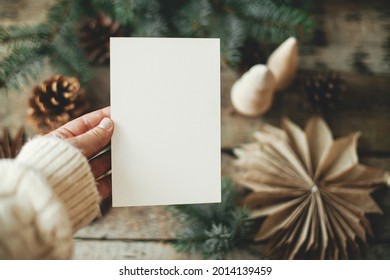 Christmas Card Mock Up. Hand Holding Empty Greeting Card On Background Of Christmas Paper Stars, Wooden Tree, Pine Branches And Cones On Rustic Wood.  Space For Text. Seasons Greetings Template