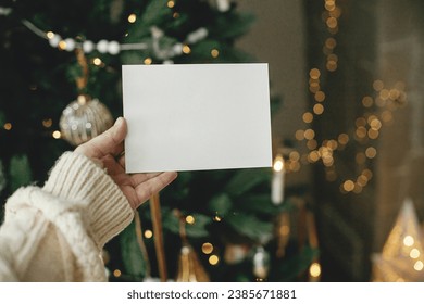 Christmas card mock up. Hand holding empty greeting card on background of stylish decorated christmas tree with golden lights. Space for text. Season greetings template and vintage ornaments - Powered by Shutterstock