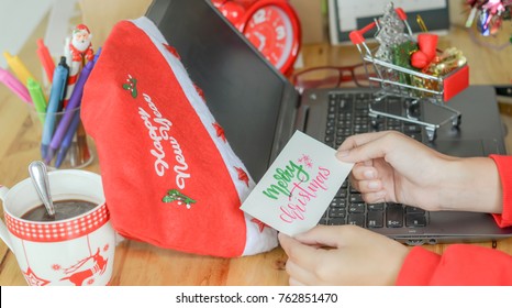 Christmas card reminder. Woman hand holding Christmas card calligraphy handwriting on white paper while working busy week at office before Xmas holiday with laptop notebook computer, red hat and props