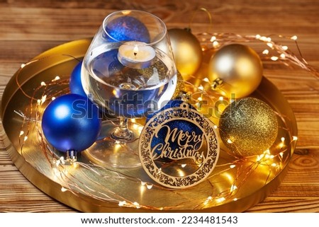Christmas card with wooden plate with Merry Christmas lettering, bright golden and blue balls, candle in the glass of water and Christmas lights