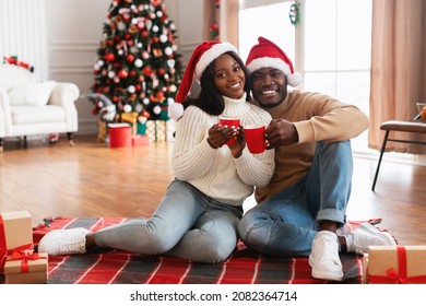 Christmas Celebration With Hot Drink. Young smiling African American husband and wife hugging, holding cups with tea or coffee in living room interior with Xmas tree, sitting on floor carpet, posing - Powered by Shutterstock