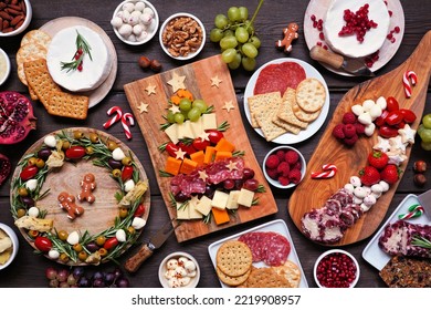 Christmas charcuterie table scene against a dark wood background. Assortment of cheese and meat appetizers. Christmas tree, wreath and candy cane arrangements.