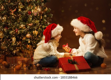 Christmas Child Girl opening Box and giving Gift Toy to Sister. Happy Children in Santa Hat next to Xmas Tree. Fairy Magic Interior, Indoor Lights - Powered by Shutterstock