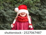 Christmas Child Girl in Santa Hat Having fun Smiling Happy Emotions on Nature Background. Funny Little Santa with Cover Eyes Waiting for Christmas Holidays.