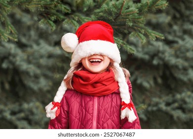 Christmas Child Girl in Santa Hat Having fun Smiling Happy Emotions on Nature Background. Funny Little Santa with Cover Eyes Waiting for Christmas Holidays. - Powered by Shutterstock