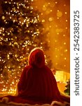 Christmas Child looking away at shining Xmas Tree. Child in Red Santa Cloak rear view in front of Christmas Lights in cozy Room. Merry Christmas and Happy Winter Holiday