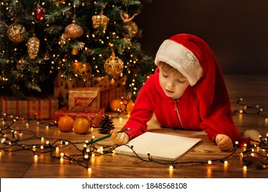 Christmas Child Write Letter To Santa Letter. Kid In Xmas Hat Writing Gift Wish List Next To Christmas Tree Over Dark Background
