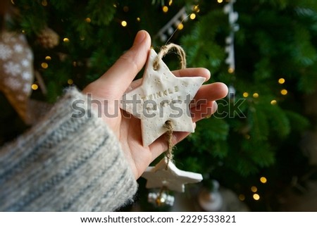 Christmas composition of female hand holding a white star ornament with a Christmas tree in background.
