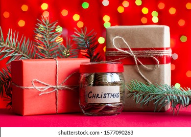 Christmas composition. Fir tree branches, coins in a glass jar with text and gift boxes on a red background with bokeh. Saving money, budget, expenses or earnings on holidays concept.