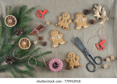 Christmas composition with gingerbread in the form of little men and spruce branches. The composition is supplemented with a root of ginger, braid, spices, scissors. Canvas background. View from above