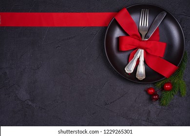 Christmas Composition With Plate, Cutlery, Pine Branches, Ribbon And Red Berries On Black Table. Winter Holidays And Festive Background. Christmas Eve Dinner, New Year Food Lunch.  Top View