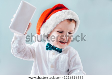 Christmas concept. Cute little baby boy in santa hat holding white gift box in hand and looks happy opening it. Plain grey backround.