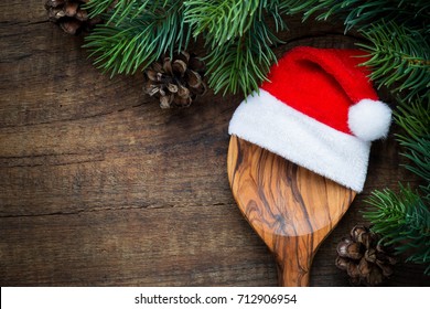 Christmas Cooking Concept - A Wooden Spoon With Santa Hat And Pine Cones On Dark Rustic Wooden Background. Overhead View With Copy Space For Your Text