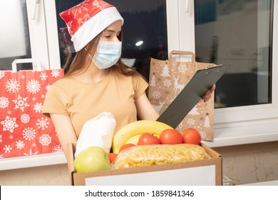 Christmas Covid Donation. A Young Woman Volunteer In Medical Mask Packing Groceries Food In A Donate Box