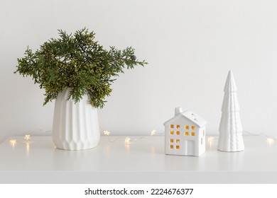 Christmas cozy winter home decor. New year interior decorations. Green fir branch in vase, decorative ceramic house and christmas tree, glowing garland lights. Stylish composition on the table.