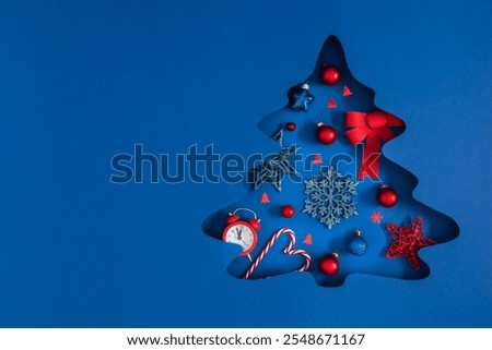 Christmas creative composition with festive Christmas tree decorations. Cut-out silhouette of fir tree and red Xmas decorations on blue paper background. Flat lay, top view