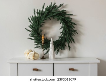 Christmas decor of the living room - a Christmas tree wreath on the wall, a brass candlestick with a burning candle, Christmas paper toys on a white chest of drawers             - Powered by Shutterstock