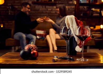 Christmas Decorations And Wine Glasses Of Red Wine On Table At Home Happy Couple Giving Christmas Gifts In Background. Winter Mood With Dark And Warm Colors.