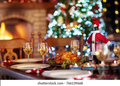 Christmas Dinner At Fireplace And Decorated Xmas Tree. Dish With Roasted Turkey, Salad And Baked Potato Served For Festive Family Meal. Wine Bottle With Santa Hat. Open Fire In Stone Oven.