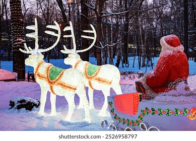 Christmas display of illuminated reindeer pulling a sleigh with Santa Claus in a snowy winter park, surrounded by trees wrapped in festive lights - Powered by Shutterstock