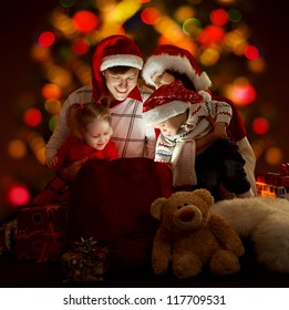 Christmas Family Of Four Persons In Red Hats Opening Lighting Bag With Gifts