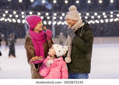 Christmas, Family And Leisure Concept - Happy Mother, Father And Daughter Eating Takeaway Pancakes At Outdoor Skating Rink In Winter