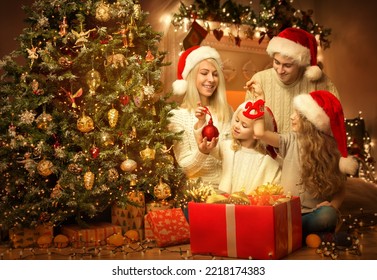 Christmas Family in Santa Hat decorating Xmas Tree. Happy Parents with Kids opening Big Red Gift Box with Presents in Dark Room Interior. Christmas Ornaments, Balls and Lights - Powered by Shutterstock