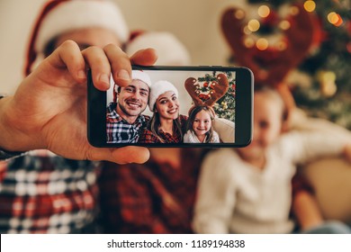 Christmas. Family. Technology. View On The Smartphone. Dad, Mom And Daughter In Santa Hats Looking At Camera And Smiling While Doing Selfie