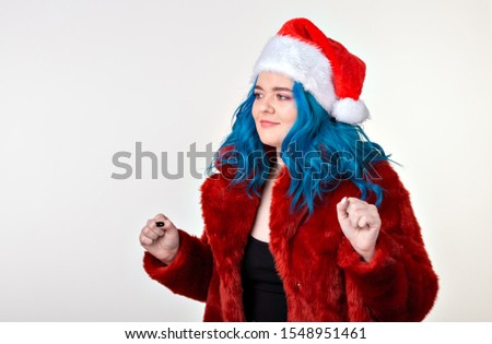 Christmas, fashion and youth - Beautiful young woman with blue hair in red fur coat and santa hat, looking at the camera and standing against a white background. Ready for party.