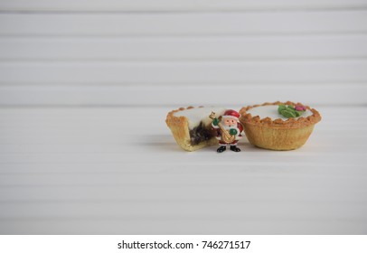 Christmas Food Photography With Mince Pie Iced With Holly Berry Decoration And Cute Small Xmas Santa Claus Father Christmas With Musical Instrument On Bright White Natural Wood Background